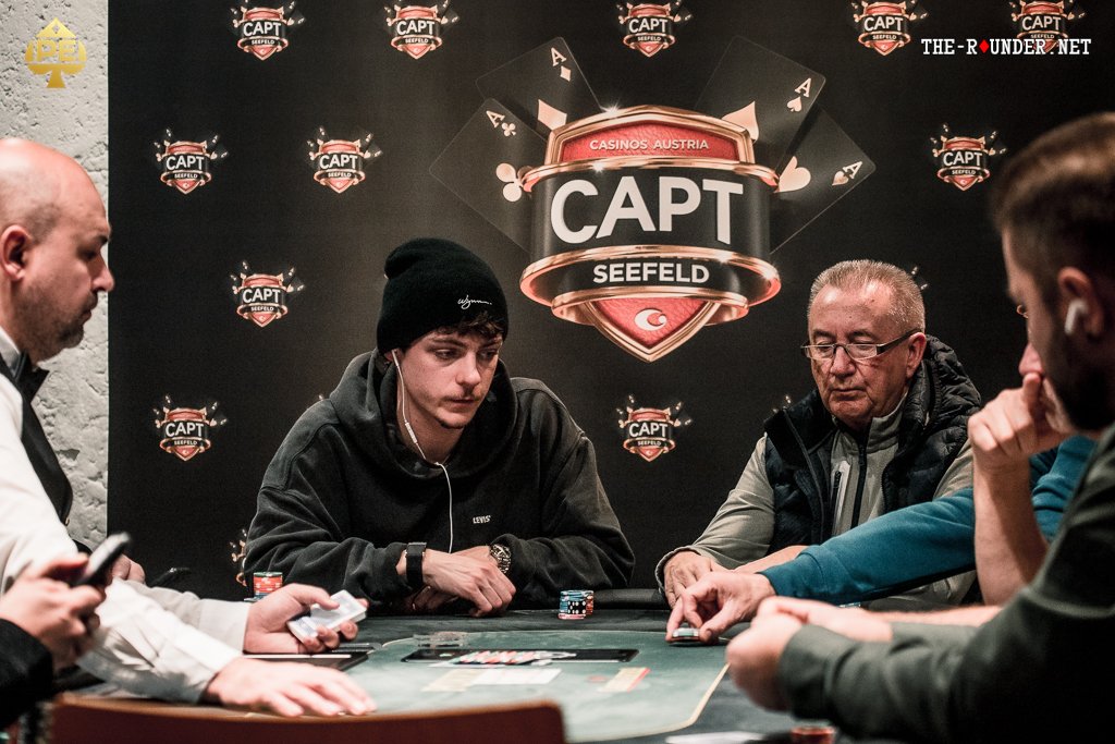 Ein Mann spielt Poker an einem Tisch, umgeben von anderen Spielern. Er hält Karten in der Hand und betrachtet konzentriert die Mitte des Tisches. Die Atmosphäre ist gespannt und die