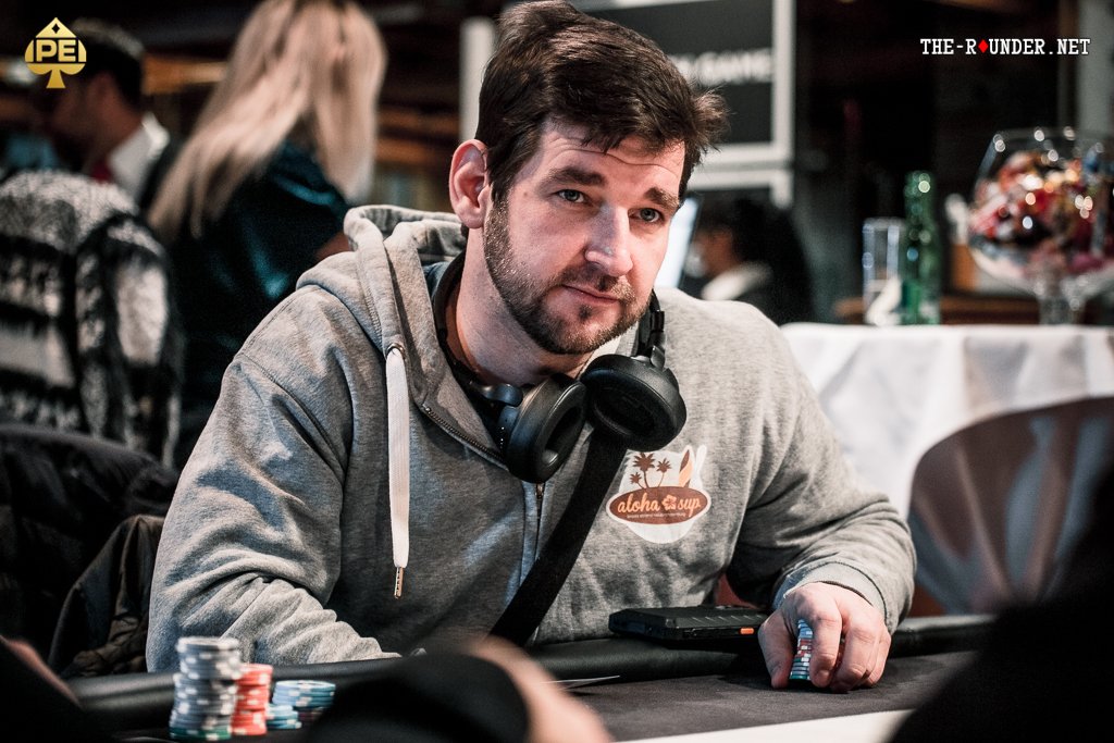 Ein Mann sitzt an einem Pokertisch und hält seine Karten in der Hand, während er konzentriert auf den Tisch schaut. Um ihn herum sind Pokerchips und Spielkarten verstreut.