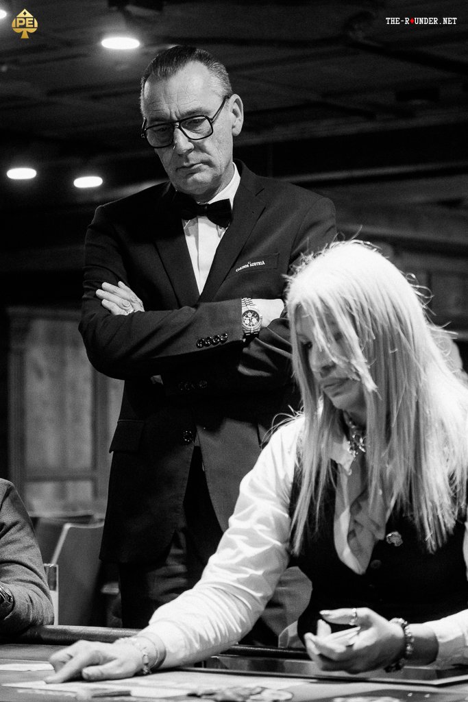 Ein Mann in einem schwarzen Anzug und einer Sonnenbrille sitzt an einem Pokertisch und hält Karten in der Hand. Er hat einen konzentrierten Blick und scheint strategisch zu überlegen. Im Hinter