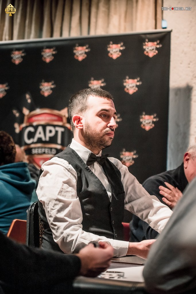Ein Mann mit einem schwarzen Anzug und einer roten Krawatte sitzt an einem Pokertisch und hält seine Spielkarten in der Hand. Gegenüber von ihm sitzt eine Person mit einem blauen Hemd und einem