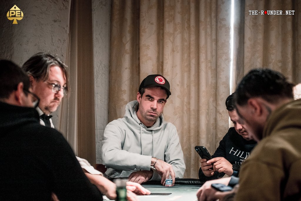 Ein Mann in einem eleganten Anzug sitzt an einem Pokertisch und hält seine Karten in der Hand. Er wirkt konzentriert und fokussiert auf das Spiel. Um ihn herum sind Pokerchips und