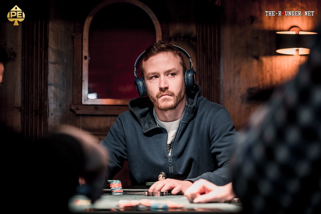 Ein Mann spielt Poker an einem Tisch mit grüner Filzdecke, umgeben von anderen Spielern. Er hält Karten in der Hand und betrachtet sie konzentriert, während Pokerchips vor ihm lie