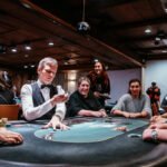 Ein Mann sitzt an einem Pokertisch und betrachtet konzentriert seine Karten, während er von anderen Spielern umgeben ist. Die Spannung ist in der Luft spürbar.