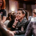 Ein Mann in einem schwarzen Anzug und einer schwarzen Sonnenbrille sitzt an einem Pokertisch und hält seine Karten in der Hand, während er konzentriert auf seine Gegner schaut.