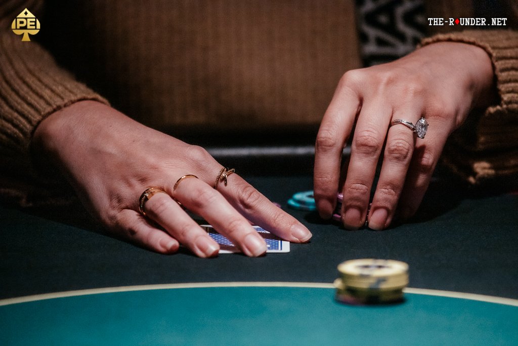 Ein Mann spielt Poker an einem Tisch in einem eleganten Casino. Er hält Karten in der Hand und betrachtet konzentriert seine Chips. Im Hintergrund sind andere Spieler und ein Croupier zu sehen.
