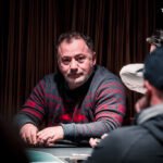 Ein professioneller Pokertisch mit grünem Filz, umgeben von Stühlen, Pokerchips und Spielkarten. Ein Spieler mischt Karten, während andere Spieler gespannt zuschauen.