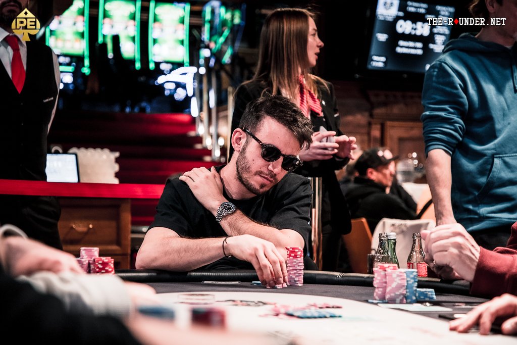Ein Mann und eine Frau sitzen an einem Pokertisch und spielen eine Partie Poker. Der Mann hält Karten in der Hand, während die Frau mit ernstem Gesichtsausdruck auf ihre eigenen Karten schaut. In