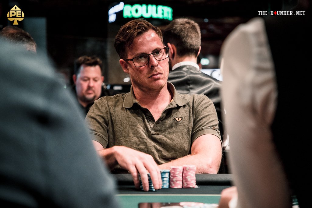Ein Mann sitzt an einem Pokertisch und hält zwei Karten in der Hand. Er schaut konzentriert auf die Mitte des Tisches, wo verschiedene Spielchips liegen. Im Hintergrund sind weitere Spieler und