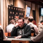 Ein Mann und eine Frau sitzen an einem Pokertisch und spielen Poker. Der Mann hält Karten in der Hand, während die Frau konzentriert auf ihre Karten schaut. Im Hintergrund sind Pokerchips und Get