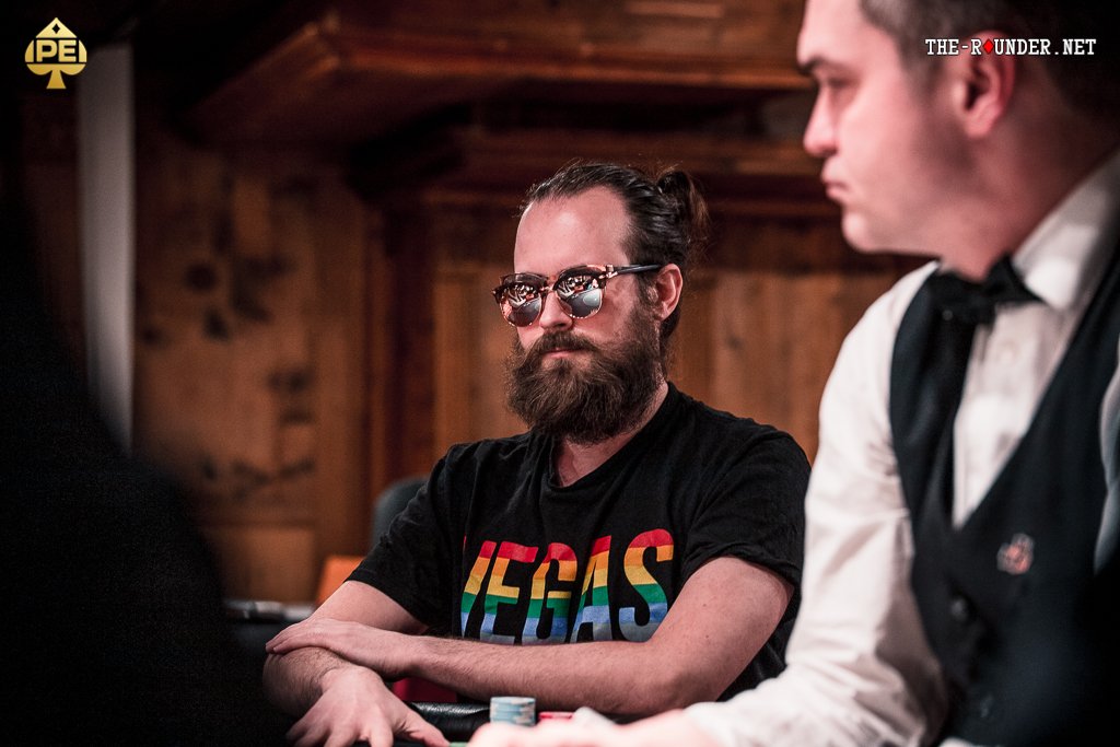 Ein Mann sitzt an einem Pokertisch mit Chips und Karten. Er betrachtet nachdenklich seine Hand, während er von anderen Spielern beobachtet wird.