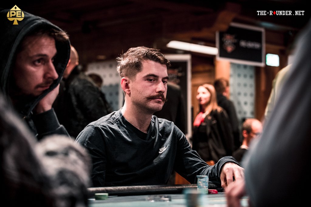 Ein Gruppenfoto von vier Personen, die an einem Pokertisch sitzen und Karten spielen. Die Spieler sind konzentriert und haben Pokerchips vor sich liegen. Ein Mann in der Mitte des Bildes trägt eine