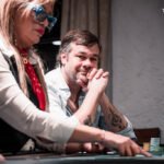 Ein Mann mit einer schwarzen Kappe und einem grauen Pullover sitzt an einem Pokertisch und betrachtet konzentriert seine Karten. Im Hintergrund sind weitere Spieler und Pokerchips zu sehen.