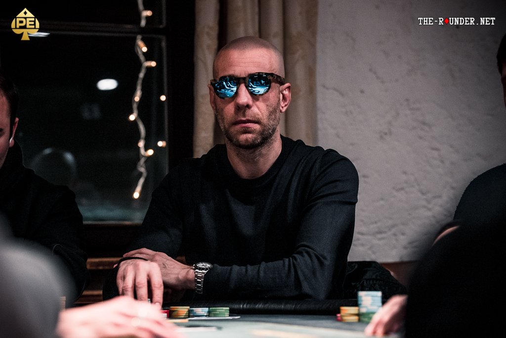 Ein bunter Pokertisch mit Spielkarten, Pokerchips und Getränken, umgeben von Menschen, die konzentriert spielen.
