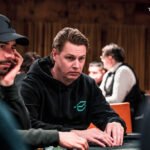 Ein Mann und eine Frau sitzen an einem Pokertisch und spielen Karten. Der Mann hat eine Hand mit Karten in der Luft, während die Frau konzentriert auf ihre Karten schaut. Im Hintergrund ist ein gr
