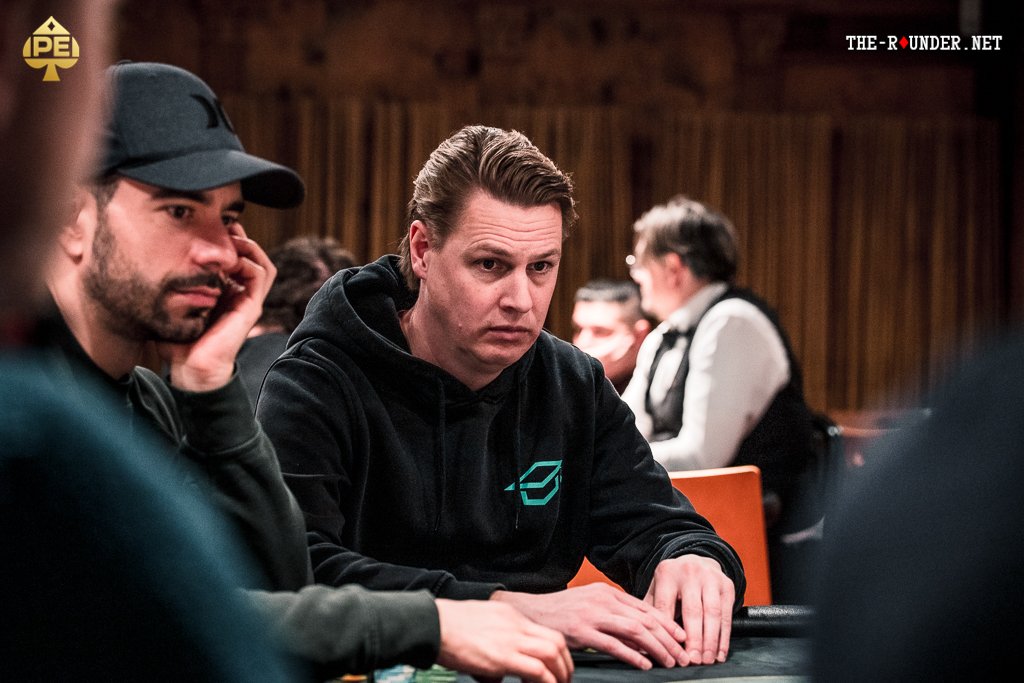 Ein Mann und eine Frau sitzen an einem Pokertisch und spielen Karten. Der Mann hat eine Hand mit Karten in der Luft, während die Frau konzentriert auf ihre Karten schaut. Im Hintergrund ist ein gr