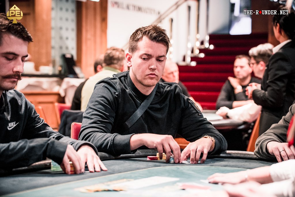 Ein Mann sitzt an einem Pokertisch und hält zwei Karten in der Hand, während er konzentriert auf seine Gegner schaut. Ein Stapel Pokerchips und eine Glücksspielkarte liegen vor ihm