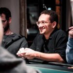 Ein Mann mit einer schwarzen Kappe und einer Sonnenbrille sitzt an einem Pokertisch und hält zwei Spielkarten in der Hand. Seine Gesichtsausdruck ist konzentriert und ernst. Im