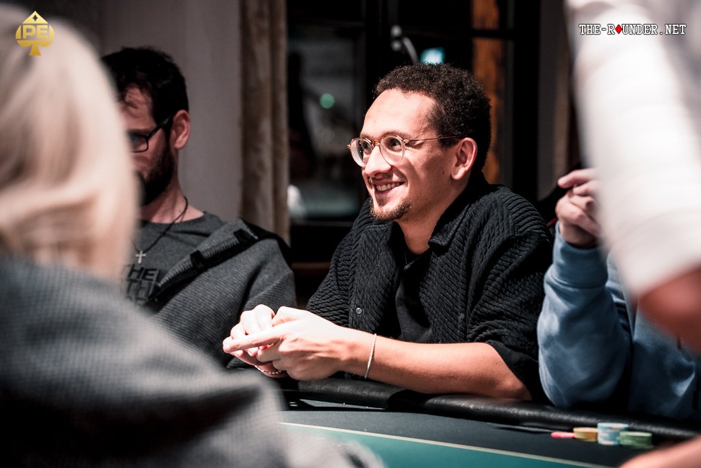 Ein Mann mit einer schwarzen Kappe und einer Sonnenbrille sitzt an einem Pokertisch und hält zwei Spielkarten in der Hand. Seine Gesichtsausdruck ist konzentriert und ernst. Im