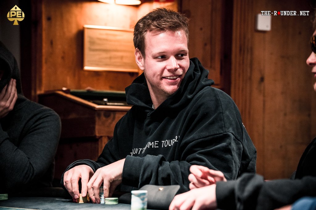 Ein Mann sitzt an einem Pokertisch und hält zwei Karten in der Hand. Er schaut konzentriert auf die Mitte des Tisches, wo Pokerchips und Spielkarten liegen. Im Hintergrund sind