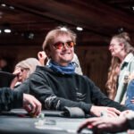 Ein Mann in einem schwarzen Anzug und einer roten Krawatte sitzt an einem Pokertisch und hält seine Karten verdeckt in der Hand, während er konzentriert auf die Karten auf dem Tisch sch