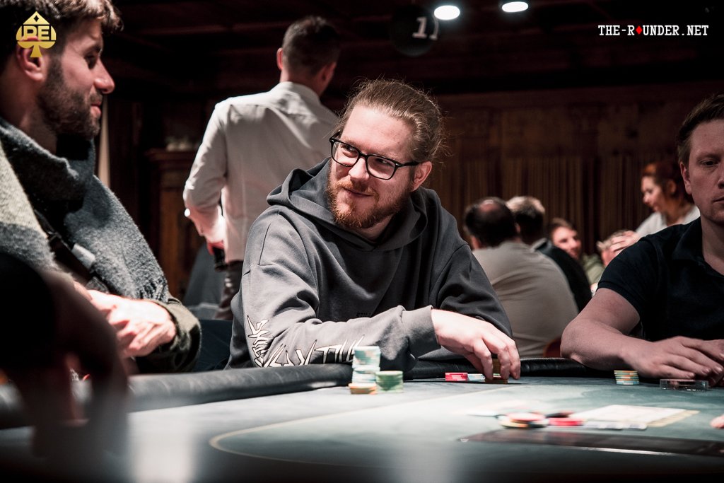 Ein Mann und eine Frau sitzen an einem Pokertisch und spielen Texas Hold'em. Der Mann hat eine Hand mit Karten und Chips vor sich liegen, während die Frau konzentriert ihre Karten betrachtet. Im Hinter
