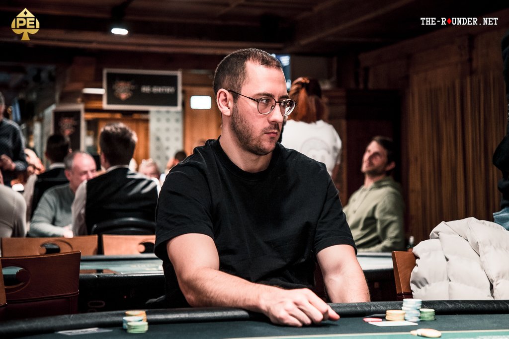 Ein Mann sitzt an einem Pokertisch und hält zwei Spielkarten in der Hand, während er konzentriert auf seine Gegner blickt. Die Tischdecke ist grün mit Pokerchips und Spielkarten