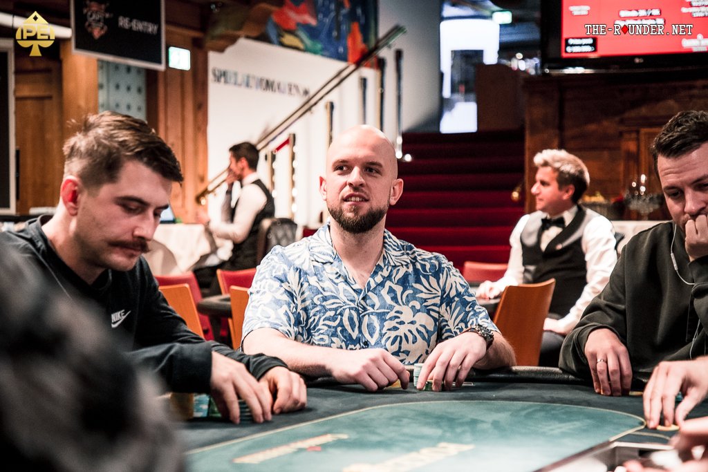 Ein professioneller Pokertisch mit grüner Filzoberfläche, umgeben von Spielern, die Karten halten und Pokerchips stapeln. Eine Mischmaschine und Getränke sind ebenfalls auf dem Tisch zu