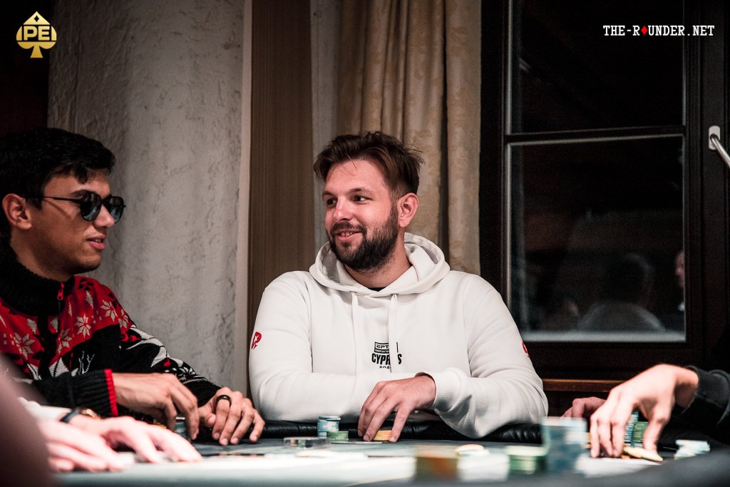 Ein grüner Pokertisch mit Spielkarten, Pokerchips und Getränken darauf, umgeben von vier Personen, die konzentriert Poker spielen.