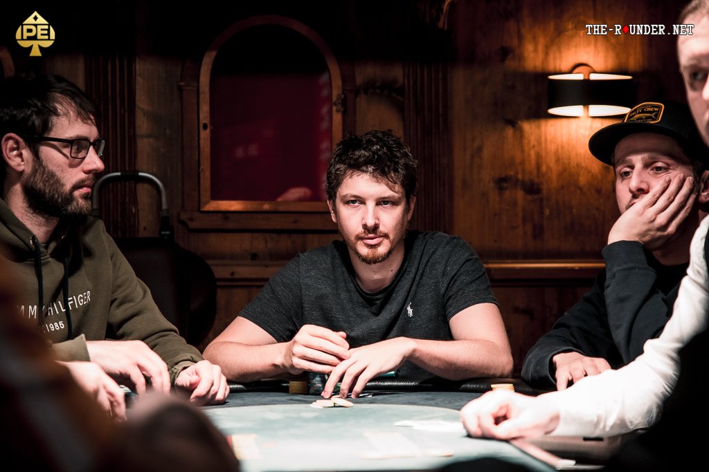 Ein Mann mit dunklen Haaren sitzt lächelnd an einem Pokertisch und hält Karten in der Hand. Er trägt ein weißes Hemd und hat eine Uhr am Handgelenk. Im Hintergrund sind