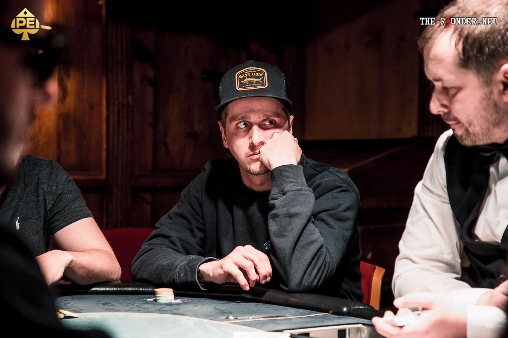 Ein Mann spielt Poker an einem Tisch mit grünem Filz. Er hält Karten in der Hand und betrachtet konzentriert seine Chips. Im Hintergrund sind weitere Spieler und eine beleuchtete Deckenle