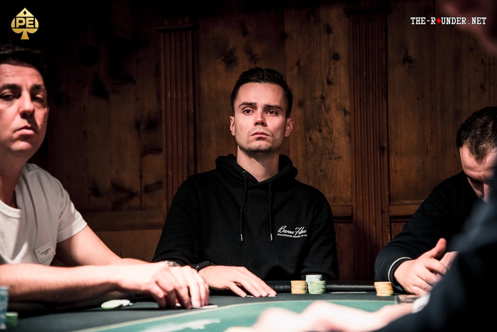 Ein Mann in einem eleganten schwarzen Anzug und einer roten Krawatte sitzt an einem Pokertisch und hält seine Spielkarten in der Hand. Um ihn herum sind Pokerchips und Gläser mit Getr