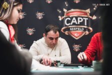Ein Mann sitzt an einem Pokertisch und hält eine Hand mit Spielkarten. Im Hintergrund sind weitere Spieler und Chips zu sehen.