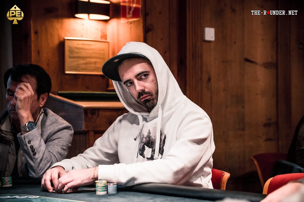 Ein Mann sitzt an einem Pokertisch und hält zwei Spielkarten in der Hand. Er schaut konzentriert auf seine Karten, während andere Spieler im Hintergrund zu sehen sind. Die Szene spielt sich in