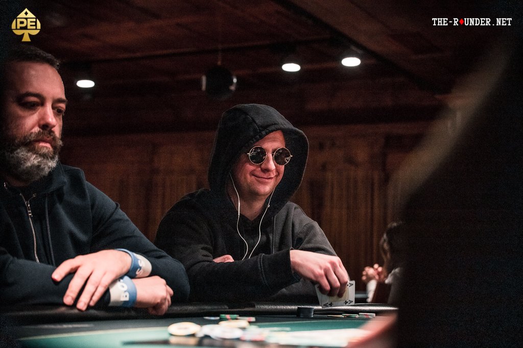 Ein Mann in einem dunkelblauen Anzug und einer weißen Hemdweste sitzt an einem Pokertisch und hält seine Karten in der Hand, während er konzentriert auf die Spielkarten auf dem Tisch