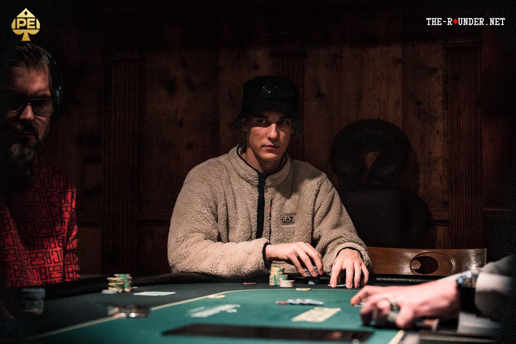 Ein Mann sitzt an einem Pokertisch und hält eine Hand mit Spielkarten. Er trägt eine dunkle Brille und konzentriert sich auf das Spiel. Andere Spieler sind im Hintergrund zu sehen.
