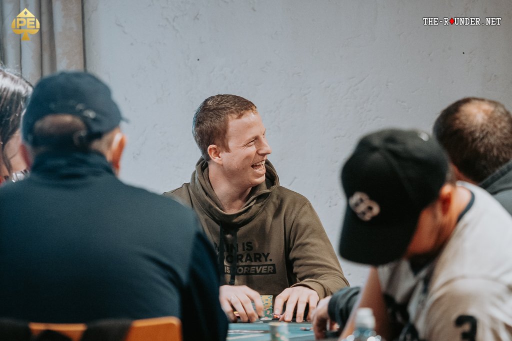 Ein Tisch mit grünem Pokertuch, auf dem Spielkarten, Pokerchips und Getränke platziert sind, umgeben von vier Personen, die konzentriert Poker spielen.