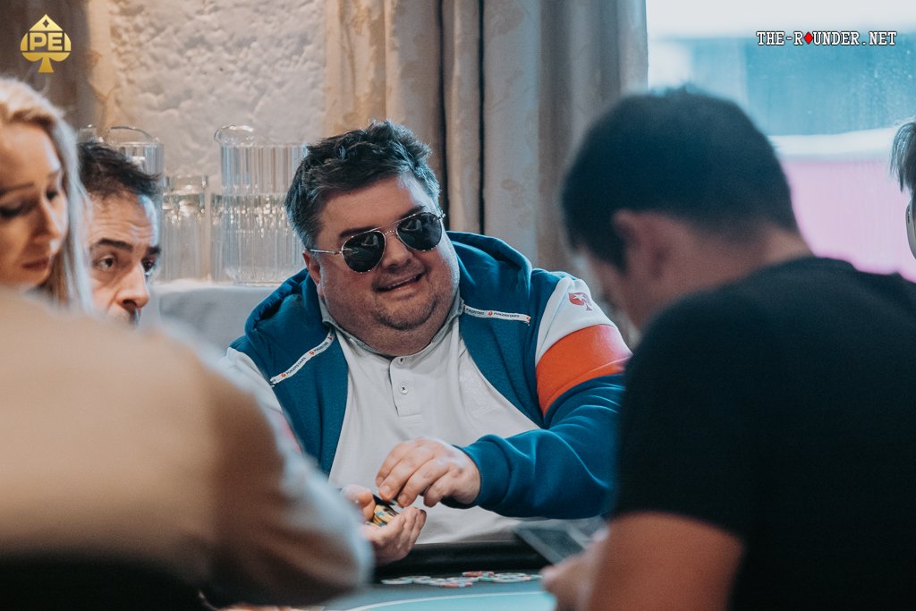 Ein Mann in einem eleganten Anzug sitzt an einem Pokertisch und hält Karten in der Hand, während er konzentriert auf seine Gegner blickt.