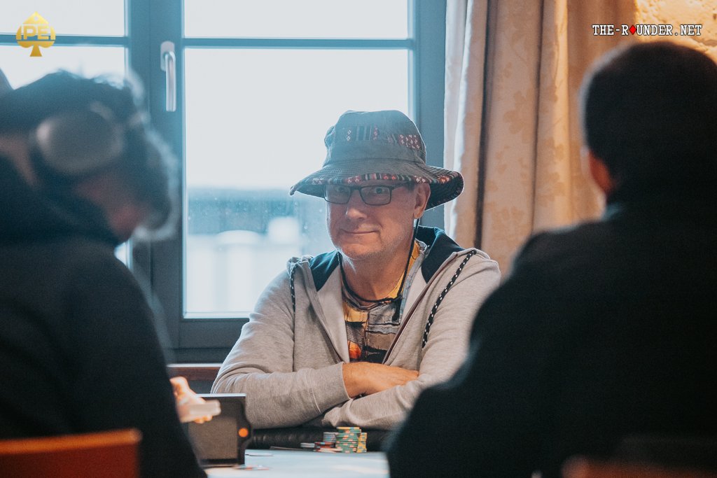 Ein Mann und eine Frau sitzen an einem Pokertisch und halten ihre Spielkarten. Der Mann hat eine ernste Miene, während die Frau lächelt. Im Hintergrund sind Pokerchips und Getränke zu sehen