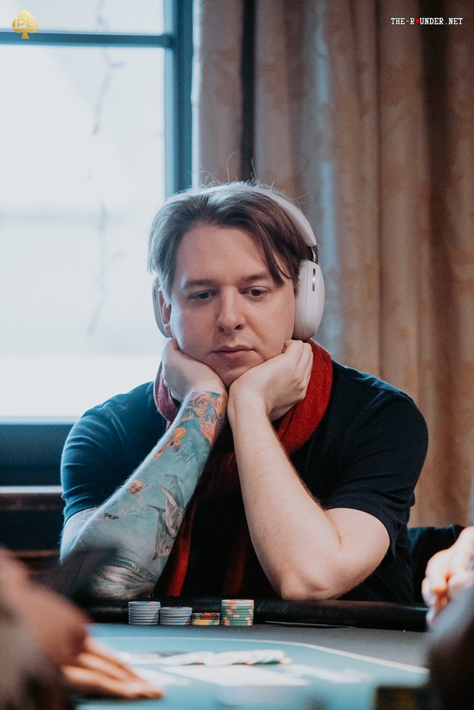 Ein Mann sitzt an einem Pokertisch und hält seine Karten in der Hand, während er konzentriert auf den Tisch schaut. Um ihn herum sind Pokerchips und ein Stapel Spielkarten zu sehen.