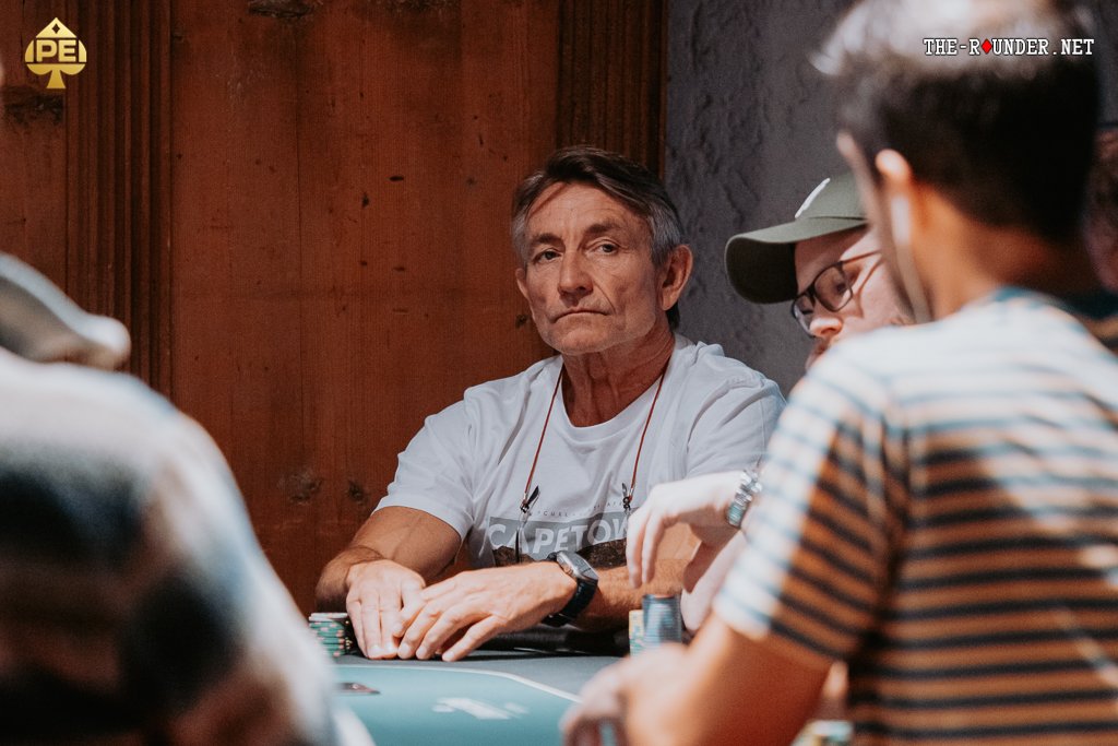 Ein Mann hält eine Hand mit fünf Spielkarten, die auf einem grünen Pokertisch liegen. Er trägt eine schwarze Kappe und konzentriert sich auf sein Blatt. Im Hintergrund sind Poker