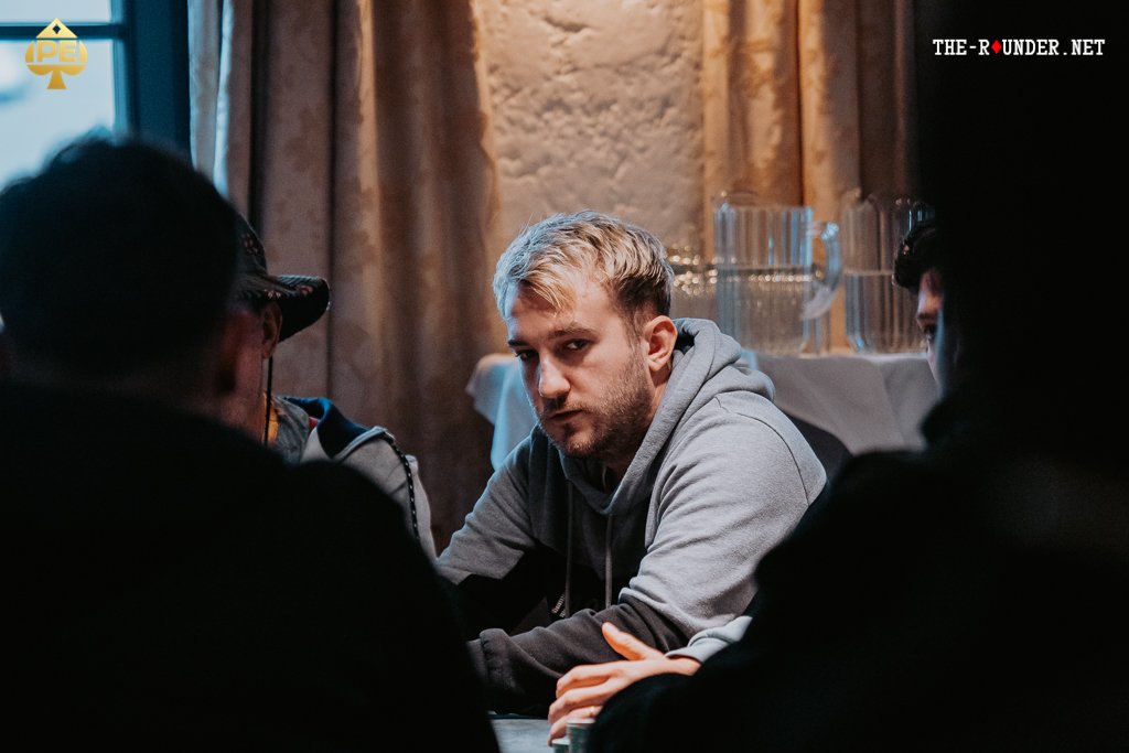 Ein Mann sitzt an einem Pokertisch und betrachtet konzentriert seine Karten, während im Hintergrund andere Spieler ebenfalls Karten halten. Die Szene strahlt eine spannende und fokussierte Atmosph