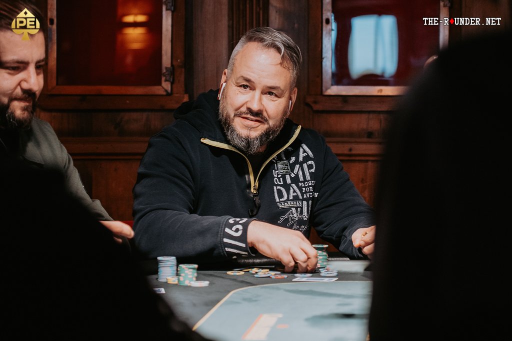 Ein Mann sitzt an einem Pokertisch, umgeben von Spielkarten und Pokerchips. Er hält seine Hand mit einem Pokerblatt in der Luft und schaut konzentriert auf den Tisch. Im Hintergrund