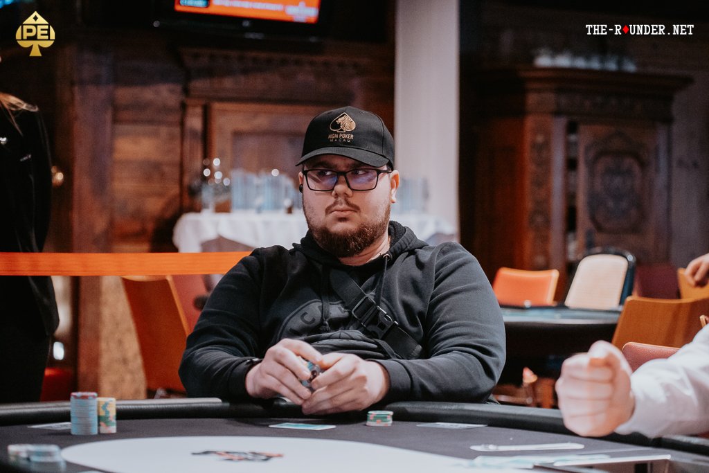 Ein Mann in einem Anzug und mit einer Krawatte sitzt an einem Pokertisch und hält zwei Spielkarten in der Hand. Er schaut konzentriert auf die Karten und wirkt ernst. Im Hinter