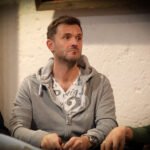 Ein Mann in einem schwarzen Anzug und einer schwarzen Sonnenbrille spielt Poker an einem eleganten Tisch mit grünen Filz. Er hält seine Karten in der Hand und betrachtet sie konzentriert