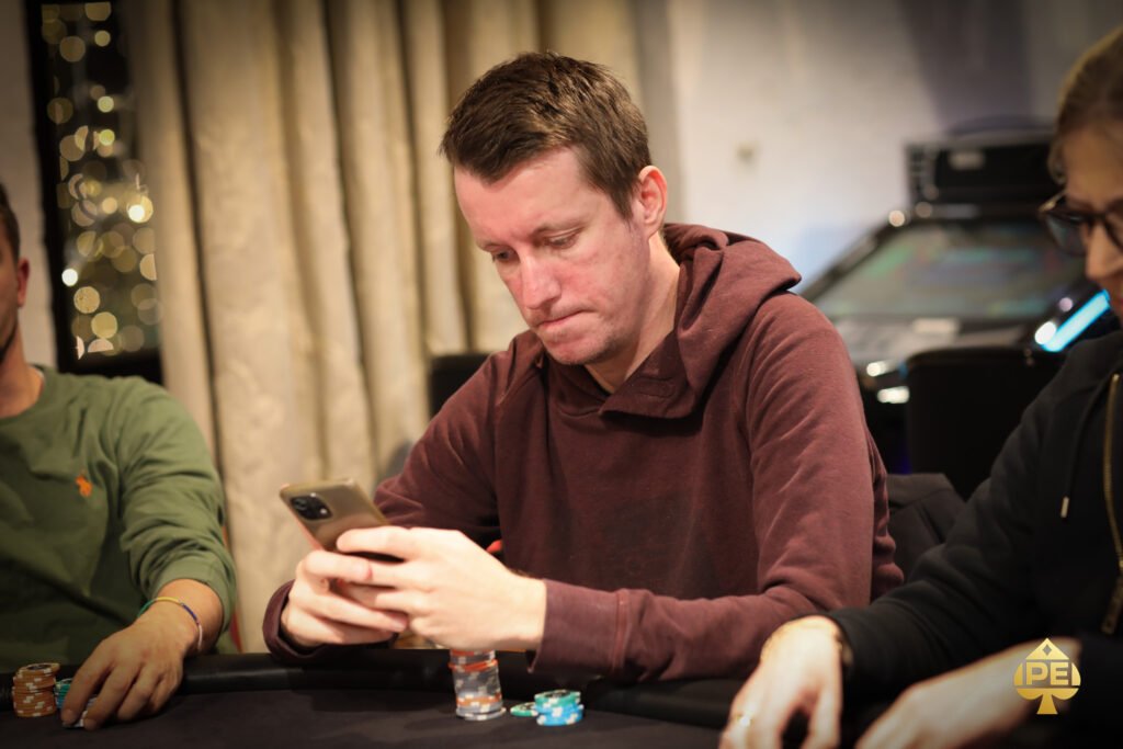 Ein Mann sitzt an einem Pokertisch, hält Karten in der Hand und schaut konzentriert auf den Tisch. In seiner Nähe sind Pokerchips gestapelt und eine Glühlampe leuchtet über