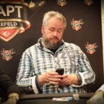 Ein Mann sitzt an einem Pokertisch und betrachtet konzentriert seine Karten. Im Hintergrund sind andere Spieler und Pokerchips zu sehen.