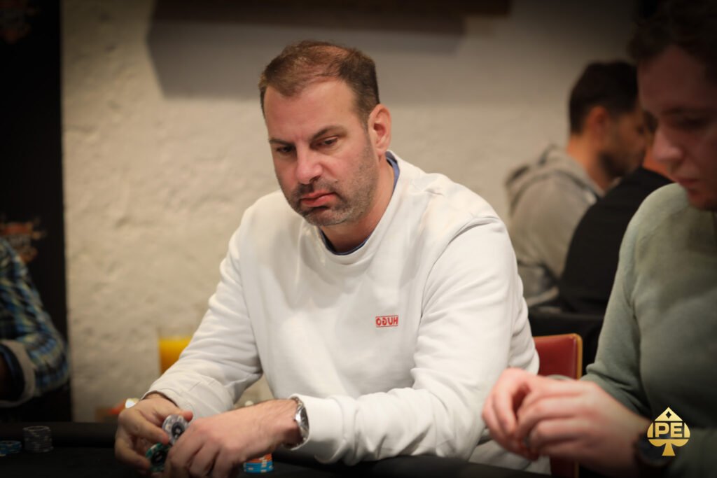 Ein Mann sitzt an einem Pokertisch und hält seine Karten in der Hand, während er konzentriert auf den Tisch schaut. Im Hintergrund sind weitere Spieler und ein Stapel Pokerchips zu sehen.