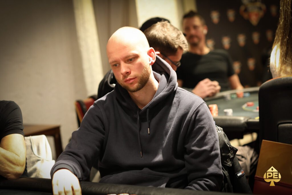 Ein Mann und eine Frau sitzen an einem Pokertisch und spielen eine Partie Poker. Der Mann hält Karten in der Hand, während die Frau konzentriert ihre Karten betrachtet. Im Hintergrund sind Pokerch