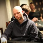 Ein Mann und eine Frau sitzen an einem Pokertisch und spielen eine Partie Poker. Der Mann hält Karten in der Hand, während die Frau konzentriert ihre Karten betrachtet. Im Hintergrund sind Pokerch