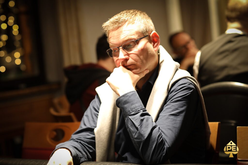 Ein Mann in einem schwarzen Anzug und einer hellen Krawatte sitzt an einem Pokertisch, während er konzentriert seine Karten betrachtet. Um ihn herum sind Pokerchips und ein Kartendeck verte